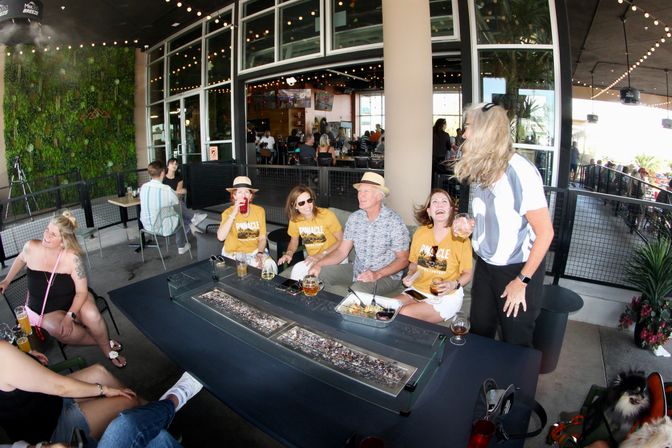 Group of friends in matching yellow shirts laughing around a long fire‑pit table on a covered outdoor restaurant patio with drinks, snacks and string lights overhead.