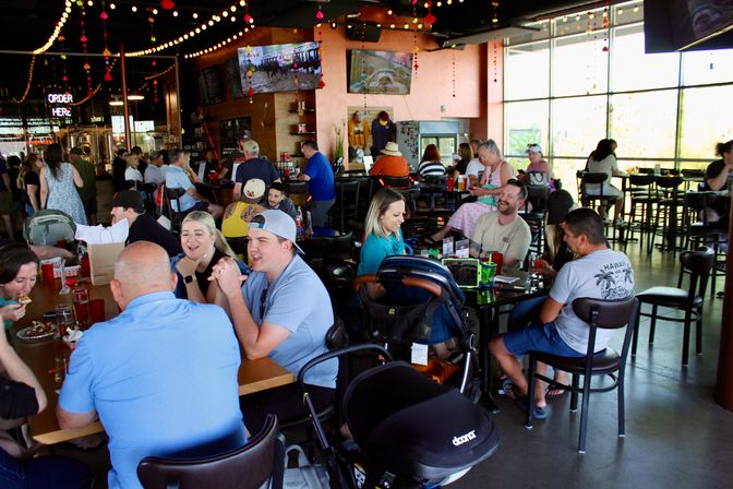 Lively family-friendly brewery taproom with groups chatting at communal tables and the bar, string lights and TVs overhead, large sunlit windows and a stroller nearby.