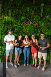 Five friends smiling and holding beer mugs in front of a lush green living wall with succulents and a geometric mountain logo on an outdoor brewery patio
