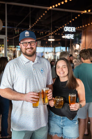 Two smiling friends in a busy brewery taproom holding pints of craft beer beneath warm string lights and a neon "ORDER HERE" sign.