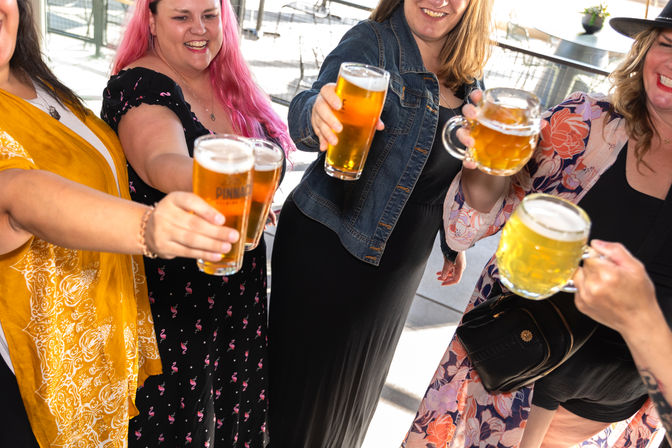 Group of friends clinking pints of beer on a sunny outdoor patio, raising glasses in a cheerful toast.