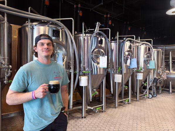 Person in a teal t‑shirt and backwards cap holding a dark beer mug inside a craft brewery, standing in front of shiny stainless‑steel conical fermenters, hoses, and brewing equipment.