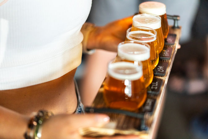 Wooden beer flight with five amber craft beer samples in tulip glasses held by a server in a busy taproom