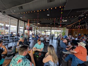 Lively craft brewery taproom with exposed ductwork and string lights, garage-style windows, high-top tables and groups of people enjoying beers