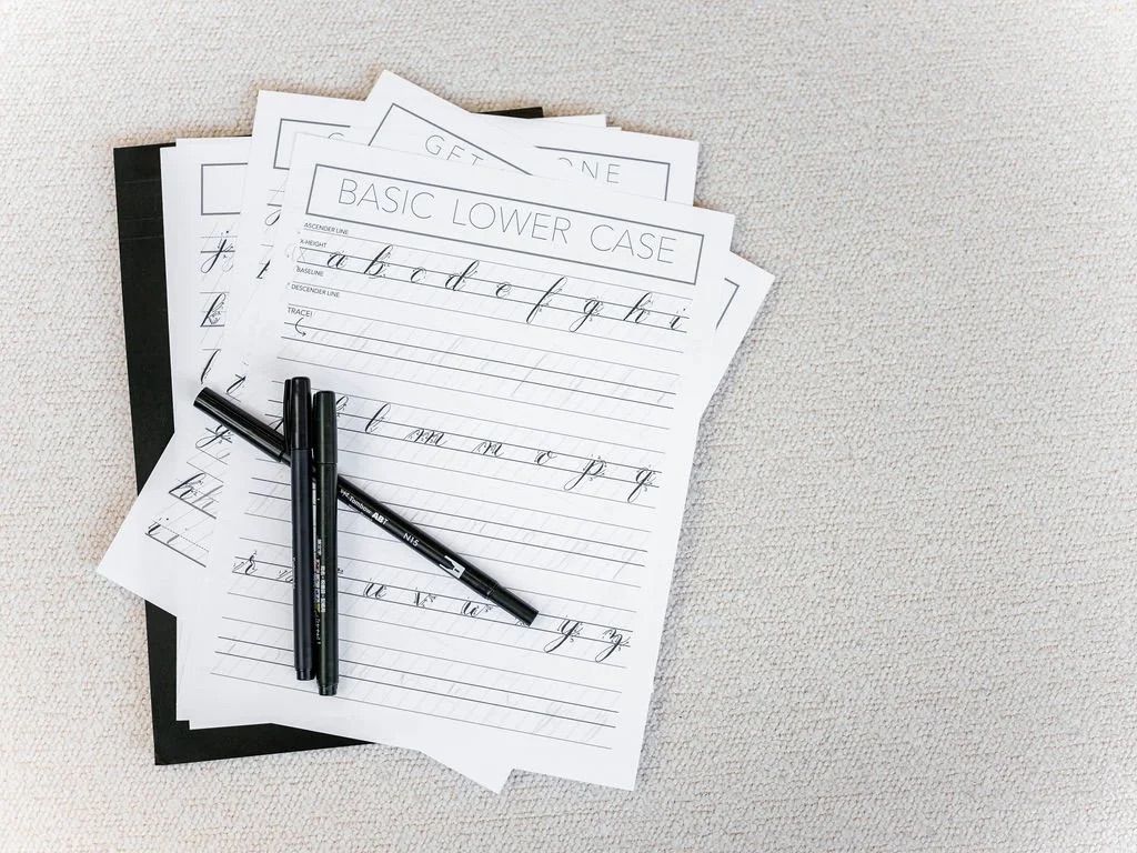Flat lay of calligraphy practice sheets labeled 'Basic Lower Case' showing cursive letters with three black brush pens on a neutral textured surface