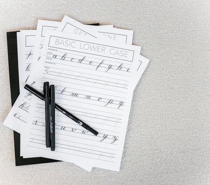 Flat lay of calligraphy practice sheets labeled 'Basic Lower Case' showing cursive letters with three black brush pens on a neutral textured surface
