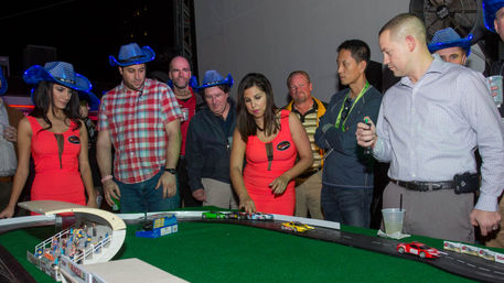 Adults gathered around a miniature slot-car racetrack at an indoor nightlife event; woman in a red dress sets cars while spectators, some wearing blue LED cowboy hats, watch.