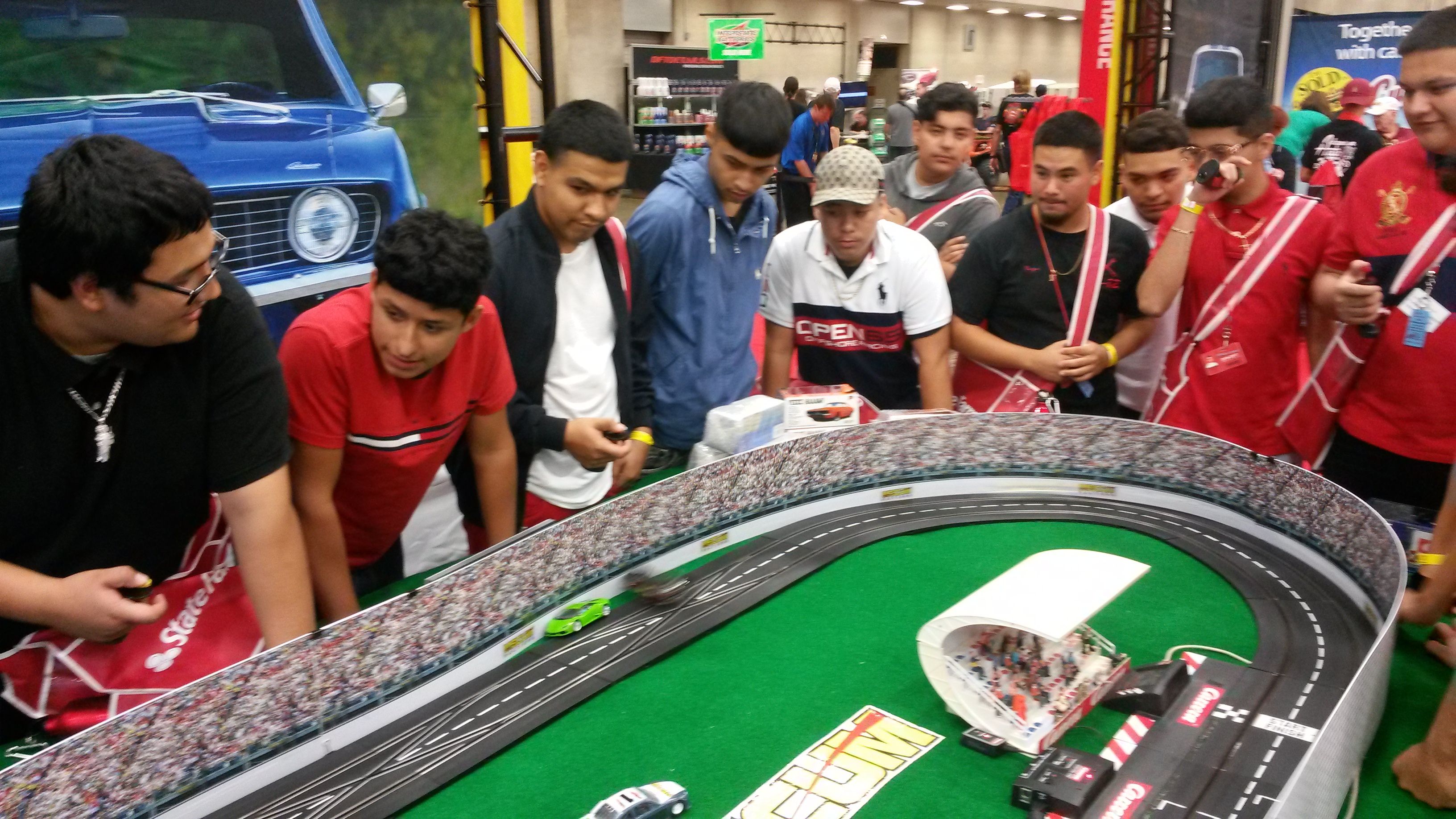 Group of teens crowded around a tabletop slot-car racetrack at an indoor hobby expo, racing miniature cars on a curved track with stadium-style scenery
