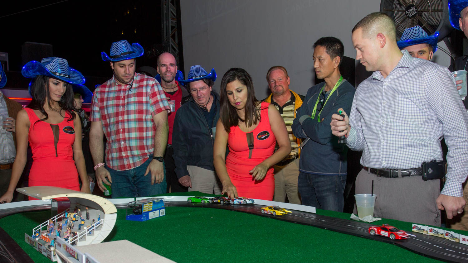 Adults gathered around a tabletop slot-car racetrack at an indoor event, women in red dresses and men—some wearing blue cowboy hats—watching and controlling toy race cars.