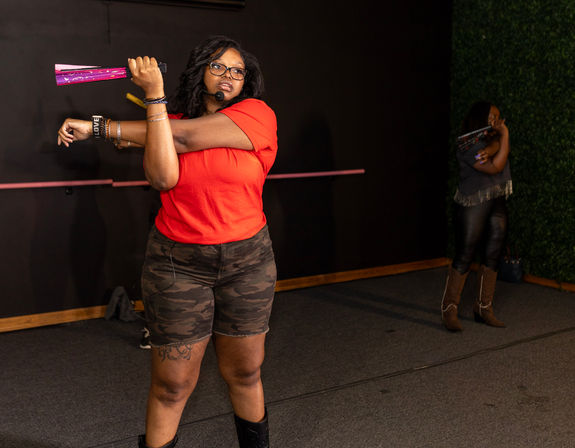 Fitness instructor in a red shirt and camo shorts stretches while holding a bright pink prop and wearing a headset mic in an indoor dance/fitness studio, with another participant near a green plant wall in the background.