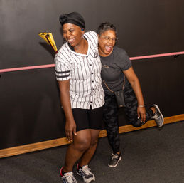Two laughing women posing in a casual indoor photo booth against a dark wall with a pink rail, one in a striped jersey and headwrap and the other in glasses, camo leggings and sneakers — candid joyful moment.