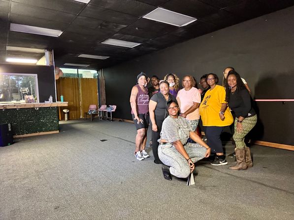 Smiling group of adult women posing in a carpeted indoor dance/fitness studio with dark walls, a barre and a small reception counter — community workout class photo.