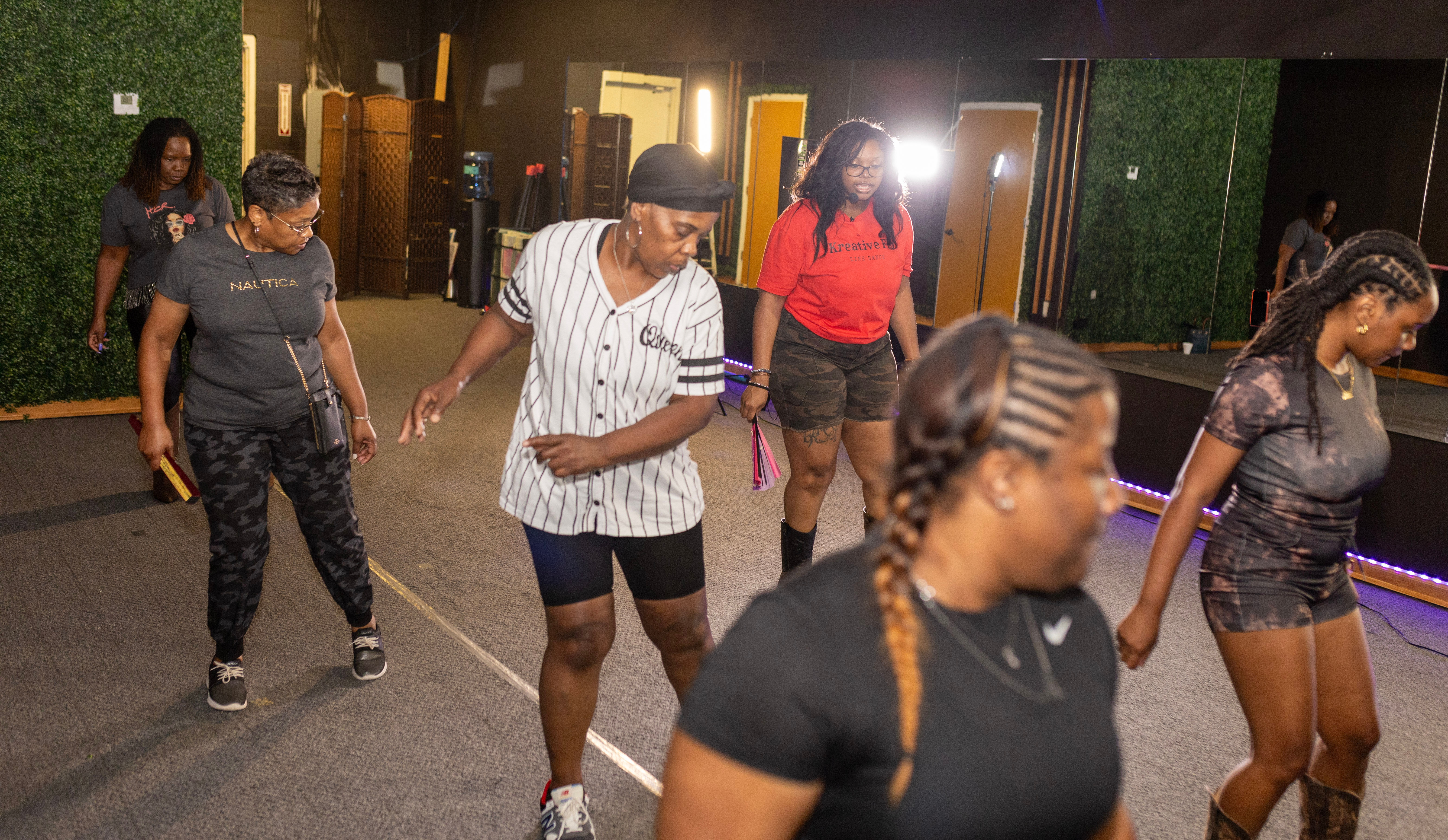 Group of women practicing a dance routine in an indoor studio with mirrors, green wall panels, and stage lights, wearing casual athletic clothing.