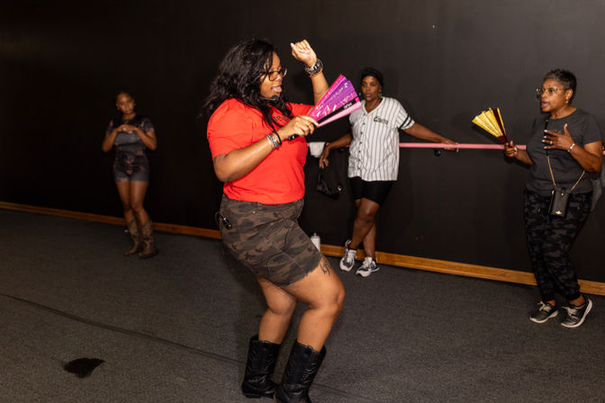 Woman in red shirt and camo shorts dancing with a purple hand fan in an indoor dance/fitness studio while three others watch and cheer, one holding a gold fan.