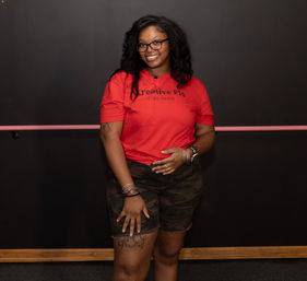 Smiling person wearing glasses, a red T-shirt with black lettering and camo shorts, posing in a dance studio in front of a pink barre and dark wall.