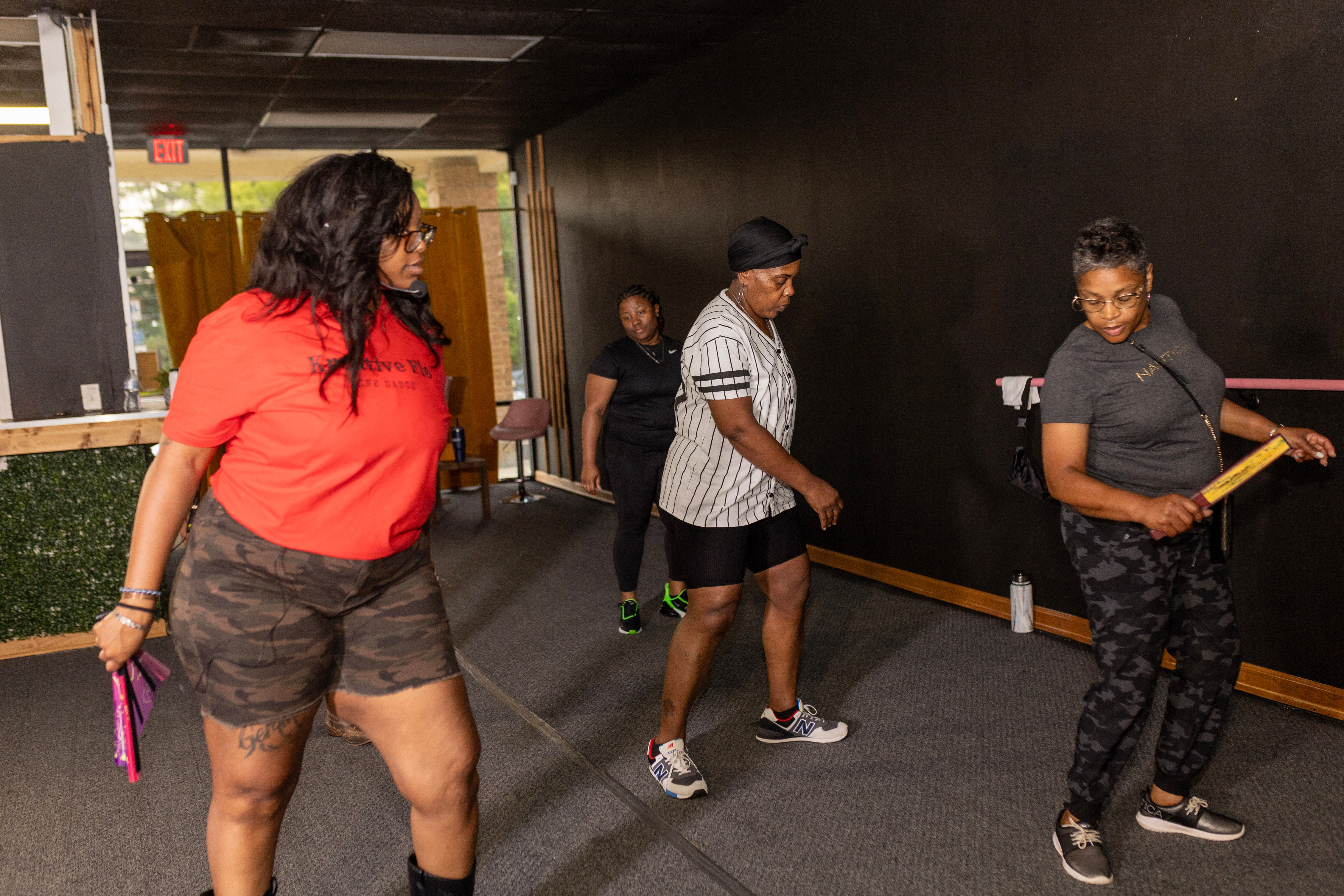 Four women in casual athletic wear practicing choreography in an indoor dance class/fitness studio with dark walls and a barre, using small hand props.
