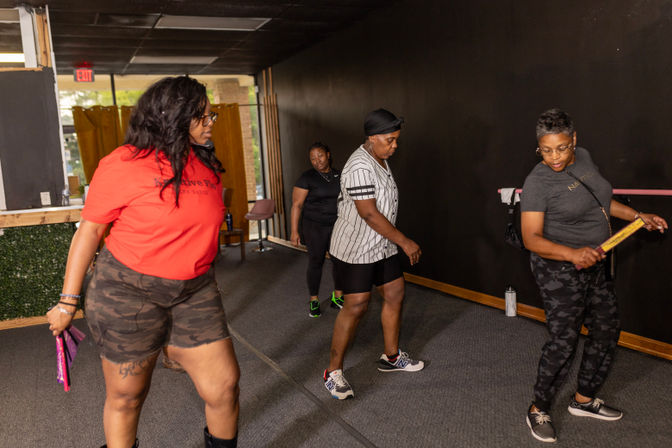 Four women in casual athletic wear practicing choreography in an indoor dance class/fitness studio with dark walls and a barre, using small hand props.
