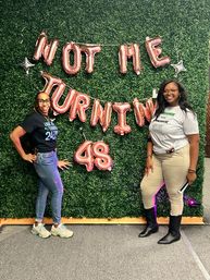 Two smiling women pose at a birthday photo backdrop with a green hedge wall and rose-gold balloons reading "Not Me Turning 48", casual outfits and boots, festive celebration vibe.