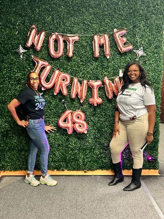 Two smiling women pose at a birthday photo backdrop with a green hedge wall and rose-gold balloons reading "Not Me Turning 48", casual outfits and boots, festive celebration vibe.