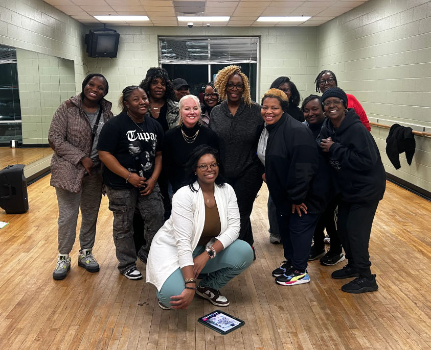 Smiling group of a dozen adults posing in a community dance studio with wooden floor, wall mirrors and a barre; one person crouches in front by a tablet on the floor while others wear casual athletic clothes and sneakers.