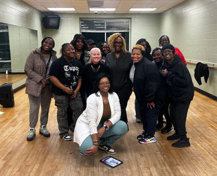 Smiling group of a dozen adults posing in a community dance studio with wooden floor, wall mirrors and a barre; one person crouches in front by a tablet on the floor while others wear casual athletic clothes and sneakers.