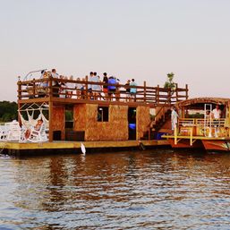 Tiki-style party barge on calm water at sunset with thatched walls, upper-deck crowd, hammocks and lounge chairs