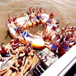 Sunny lake boat party with a large circular inflatable raft and wooden dock, dozens of people in swimwear lounging, cheering and holding drinks over brown water.