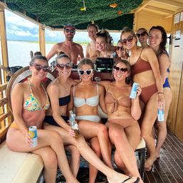 Smiling group of friends in swimsuits and sunglasses posing on a covered wooden boat, holding cans and bottles with water and shoreline visible in the background.