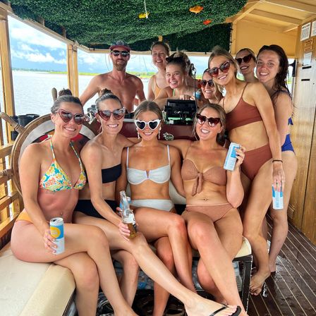 Smiling group of friends in swimsuits and sunglasses posing on a covered wooden boat, holding cans and bottles with water and shoreline visible in the background.