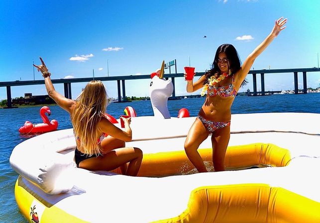 Two women in bikinis enjoying a sunny boat party on a large yellow inflatable raft with unicorn and flamingo floats near a bridge over blue water