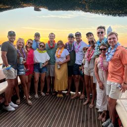 Group of friends on a sunset boat cruise wearing colorful Hawaiian leis and casual summer outfits, smiling on a wooden deck with ocean and golden sky in the background.