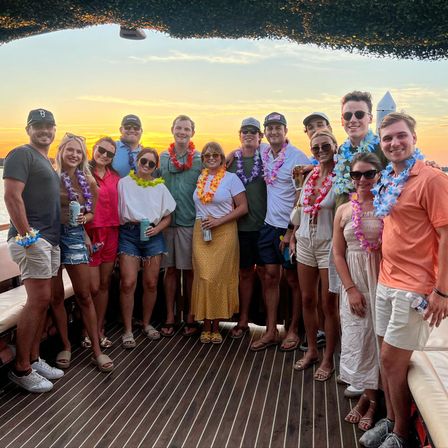 Group of friends on a sunset boat cruise wearing colorful Hawaiian leis and casual summer outfits, smiling on a wooden deck with ocean and golden sky in the background.
