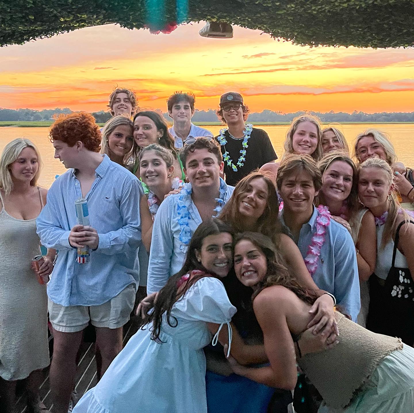 Smiling group of friends on a lakeside dock at sunset, enjoying a summer waterfront party with leis and casual outfits under a colorful orange-pink sky.