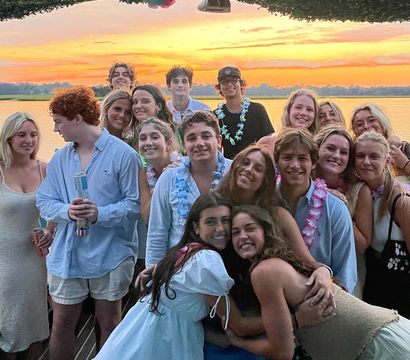 Smiling group of friends on a lakeside dock at sunset, enjoying a summer waterfront party with leis and casual outfits under a colorful orange-pink sky.
