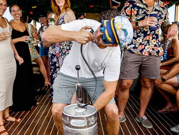 Lively boat party on a wooden deck — a man in a tropical cap taps a keg while friends in Hawaiian shirts and summer dresses laugh and hold drinks.