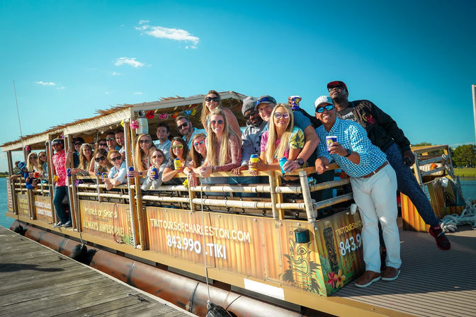 Smiling group leaning over the rail of a tiki-style tour boat at a sunny waterfront dock, holding drinks and posing for a lively tour photo.