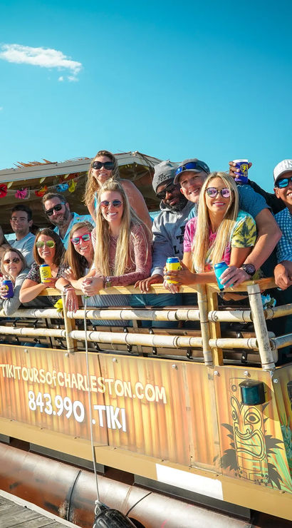 Smiling group leaning over the rail of a tiki-style tour boat at a sunny waterfront dock, holding drinks and posing for a lively tour photo.