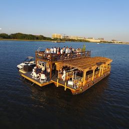 Sunlit floating tiki bar on a two-level party barge with a thatched roof and rooftop deck full of people cruising on calm water with a distant city skyline.