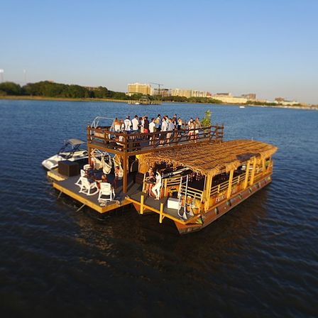 Sunlit floating tiki bar on a two-level party barge with a thatched roof and rooftop deck full of people cruising on calm water with a distant city skyline.