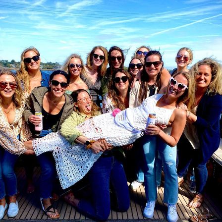 Smiling group of women on a sunny coastal boat deck lifting a woman in a white lace dress and sash for a bachelorette celebration, many wearing sunglasses and holding canned drinks