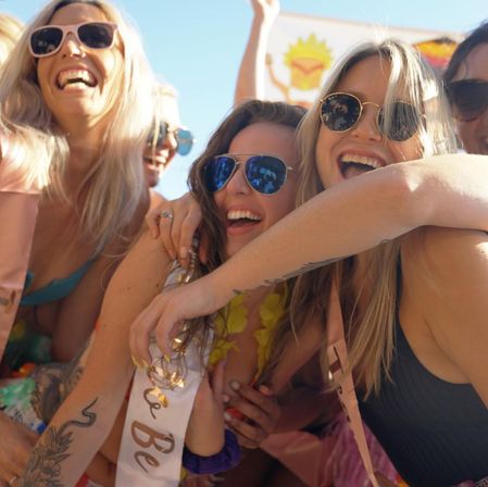 Laughing women in sunglasses hugging at a sunny outdoor summer bachelorette party, colorful swimsuits and sashes.