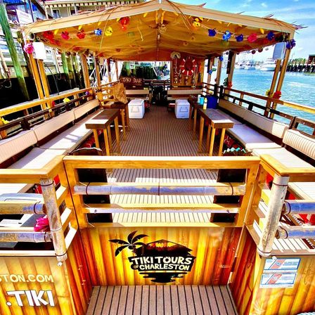 Open-air tiki-style tour boat docked at a Charleston harbor with wooden benches and tables, colorful flower garlands under a shaded canopy, and marina water views.