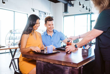 Smiling couple seated at a wooden counter in a bright, sunlit modern café, being handed a drink bottle by a staff member