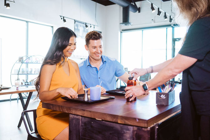 Smiling couple seated at a wooden counter in a bright, sunlit modern café, being handed a drink bottle by a staff member