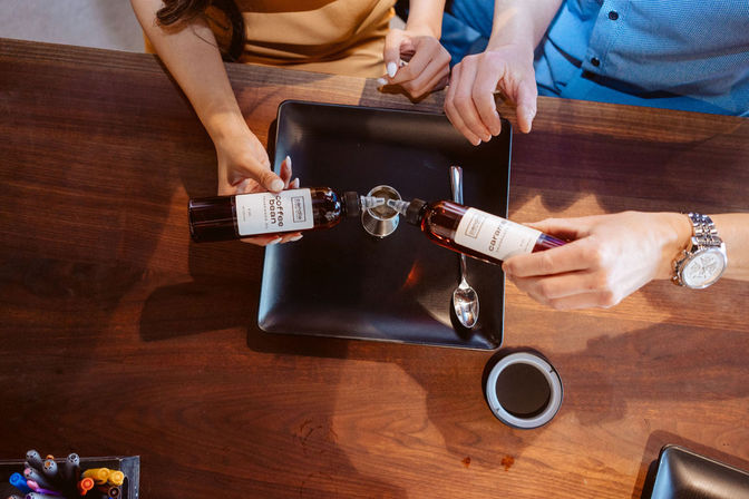 Overhead shot of two people pouring flavored syrup bottles into a small glass on a black square plate, a spoon on the plate, a wristwatch visible, and a coffee cup lid on a wooden table.