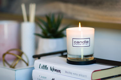 Lit white glass candle on a coaster beside a potted succulent and decorative accents, sitting atop stacked books including 'Little Women' — cozy reading nook decor