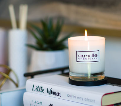 Lit white glass candle on a coaster beside a potted succulent and decorative accents, sitting atop stacked books including 'Little Women' — cozy reading nook decor