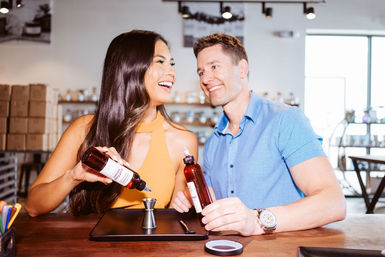Cheerful pair pouring amber liquid from labeled bottles into a metal jigger on a wooden counter inside a bright artisan apothecary-style boutique