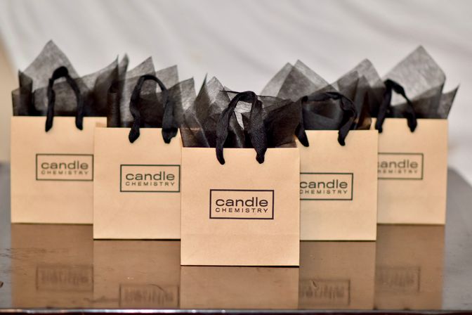 Five kraft gift bags with black tissue paper and ribbon handles arranged on a polished wooden table — boutique candle gift favors ready for an event.