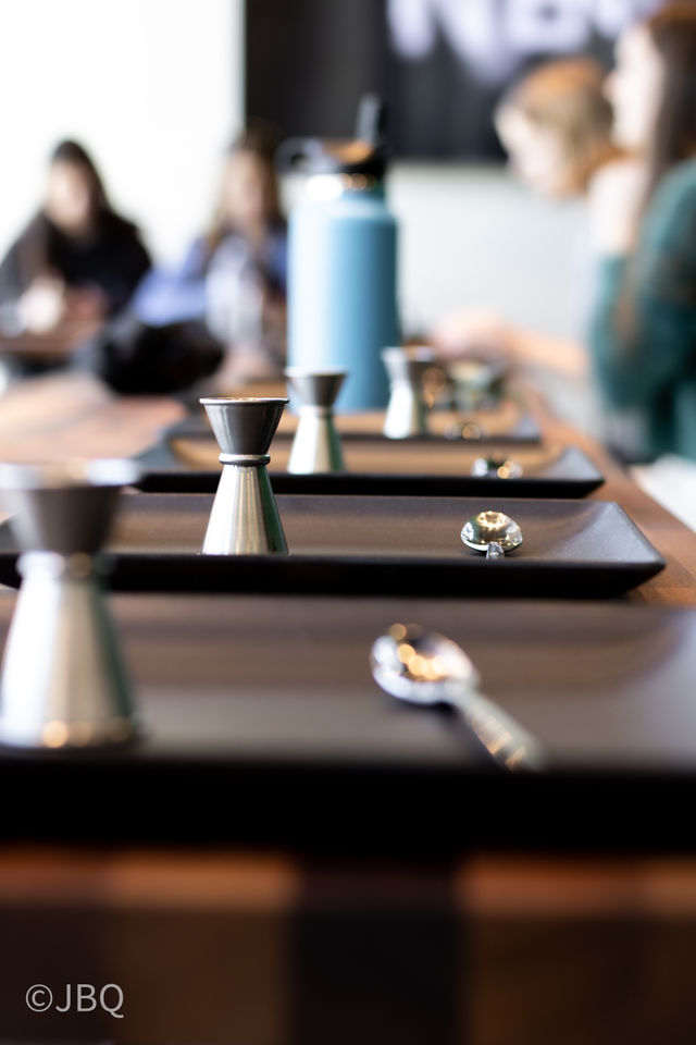 Close-up of stainless-steel jiggers and spoons lined on black trays on a wooden cafe counter, blurred customers and a blue bottle in the background.